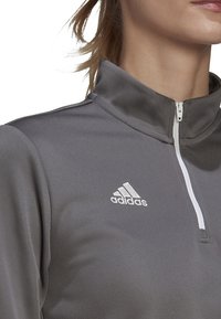 Maglia sportiva grigia a manica lunga con zip a quarto di altezza, realizzata in materiale liscio, presenta un colletto alto, una zip bianca e il logo Adidas sul lato sinistro del petto.
