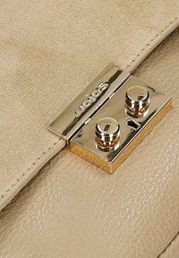 Fermoir en métal doré avec deux boutons cylindriques sur le rabat d’un sac à main en cuir beige texturé et daim avec le logo "Sephora".