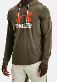 Homme portant un hoodie Under Armour vert olive avec un grand logo orange et des cordons blancs, associé à un pantalon de couleur claire.