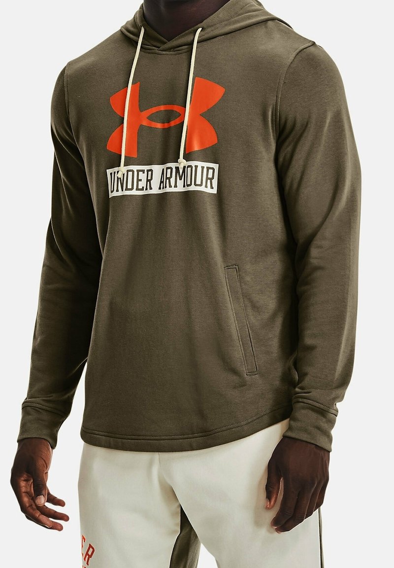Homme portant un hoodie Under Armour vert olive avec un grand logo orange et des cordons blancs, associé à un pantalon de couleur claire.