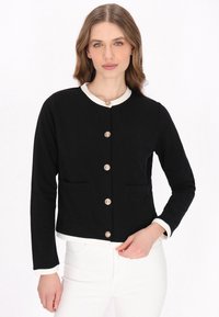 Jachetă neagră cu guler rotund, detalii crem, patru buzunare frontale și butoane decorative. Realizată dintr-un material moale cu o croială ajustată.
