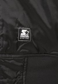 Giacca Black Starter Black Label con tessuto testurizzato, caratterizzata da un patch logo prominente. Il design include una superficie liscia e cuciture visibili.