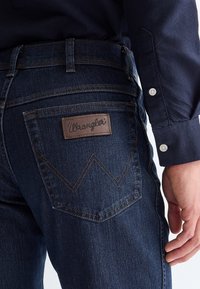 Gros plan sur la hanche d'une personne portant un jean Wrangler bleu foncé et une chemise bleu marine à manches longues avec des boutons blancs aux poignets.