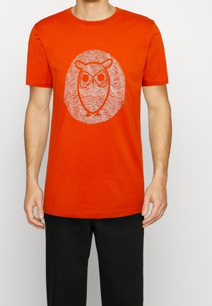 Orange bomulds T-shirt med et hvidt grafik af en ugle inden for bølgede linjer, rundt design, korte ærmer og rund hals.