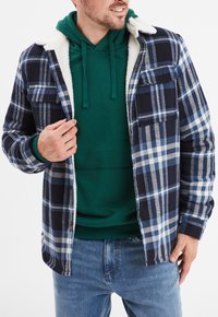 Camicia in flanella a quadri nei colori navy, blu e bianco con collo foderato in sherpa. Indossata sopra un hoodie verde, abbinata a jeans blu.