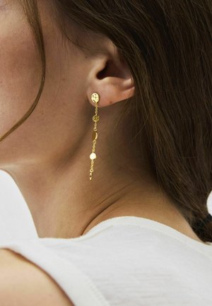 Nahaufnahme vom Ohr einer Frau, die einen langen goldenen Kettenohrring mit kleinen runden Scheiben trägt, vor braunem Haar und weißem Oberteil.