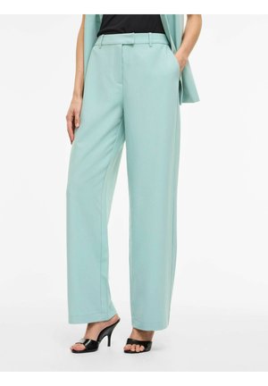 Femeie purtând pantaloni largi albastru deschis cu buzunare laterale, asortați cu sandale negre cu toc și deschise în față și o jachetă fără mâneci asortată.