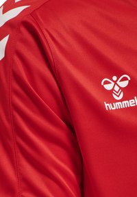 Rode sportshirtstof met wit Hummel-logo met een gestileerde hommel en chevron-ontwerp op de schouder.