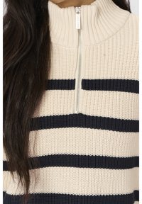 Pull tricoté rayé beige et noir avec un col haut et une demi-fermeture éclair, porté par une personne aux longs cheveux foncés.