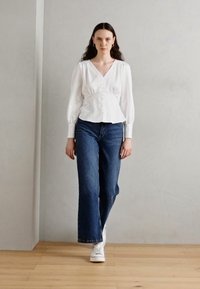Blouse blanche à manches longues avec un décolleté en V et des détails de boutons, associée à un jean en denim bleu taille haute évasé et des baskets blanches.