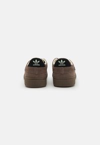 Sneaker in suede marrone con accenti in tessuto nero, dotati di una suola in gomma con texture e lacci bianchi. Logo Adidas sul tallone e sulla linguetta.