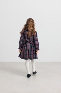 Καρό φόρεμα με μακριά μανίκια και ζώνη στη μέση, με σκούρους μπλε, κόκκινους και πράσινους σχεδιασμούς. Λευκές ριπτές καλσόν και μαύρα παπούτσια.