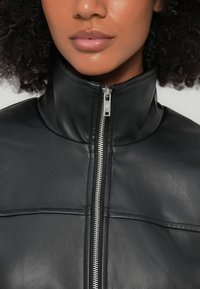 Jachetă neagră din piele cu guler înalt, fermoar frontal și detalii cusute pe panouri. Textură netedă cu un design minimalist.