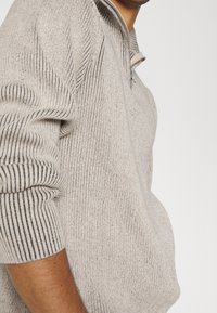 Maglione grigio chiaro a costine con collo con cerniera e maniche lunghe. Presenta un tessuto strutturato con macchie scure e una vestibilità comoda.