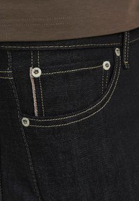 Jeans en denim bleu foncé avec des coutures jaunes, arborant une poche avant, des boutons métalliques et une bande d'accent blanche et rouge.
