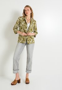 Blazer vert olive avec imprimé de feuilles blanches, manches trois-quarts, associé à un jean gris clair retroussé et des mules en plateforme marron.