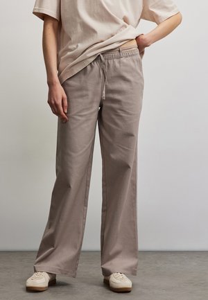 Person trägt beige, weite Hose mit Kordelzug, hellbeiges T-Shirt teilweise in die Hose gesteckt und cremeweiße Sneakers, steht auf grauem Boden.