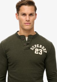 Olivengrøn langærmet henley-skjorte med en struktureret overflade, som har en knaplukning ved halsen og et cremefarvet "SUPERDRY 23" logo på brystet.