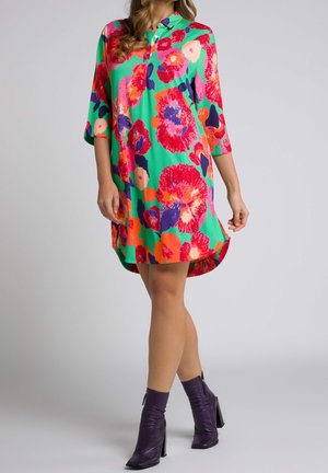 Femme portant une robe verte avec de grands motifs floraux rouges et orange et des bottines violettes, debout devant un fond clair uni.