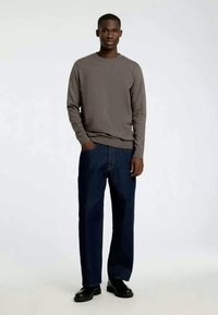 Grå langermet skjorte med rund hals, sammen med mørkeblå jeans med rette ben, iført svarte sko, stående på en nøytral bakgrunn.