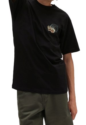 Mężczyzna w czarnej koszulce Vans z małym logo i grafiką na piersi, w oliwkowozielonych spodniach, z uniesioną ręką ponad głową.