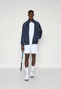 Marinblå sportjacka med vita detaljer, vit t-shirt och vita shorts; håller i ett tennisracket, bär vita sneakers och strumpor.