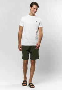 Vit bomull t-shirt med korta ärmar och en liten svart logotyp, matchad med olivgröna shorts och bruna sandaler. Enkel och avslappnad stil.