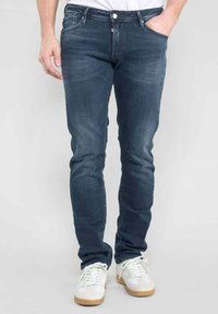 Jeans en denim bleu foncé avec une coupe slim, dotés de cinq poches et de détails légèrement décolorés. Portés avec des baskets blanches et un t-shirt uni.