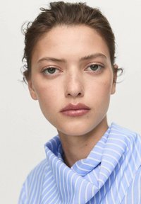 Young woman with light brown hair pulled back, wearing a blue and white striped shirt with a high collar, neutral expression, light makeup.