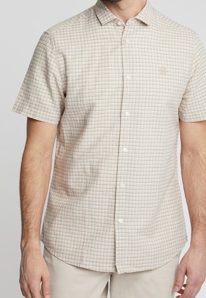 Mężczyzna w beżowo-białej kraciastej koszuli na krótki rękaw z kołnierzykiem, beżowe spodnie, na jednolitym jasnoszarym tle.