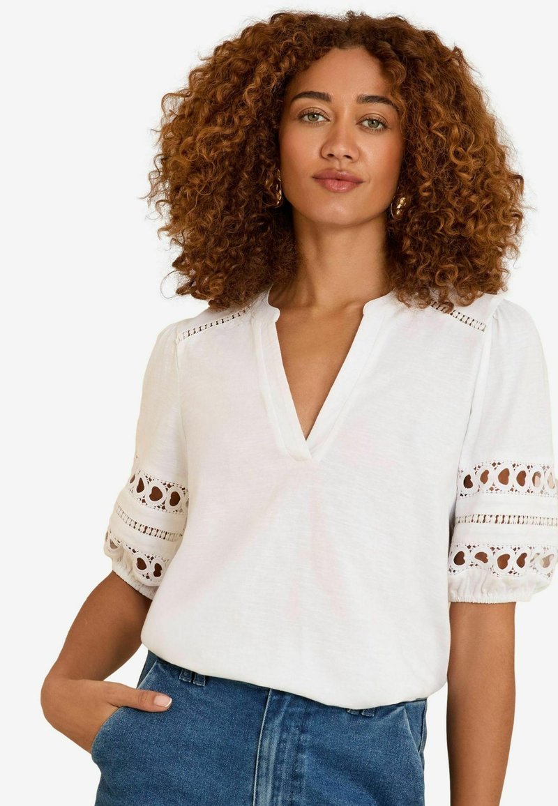 Witte blouse met korte mouwen en een V-hals, met kanten accenten op de mouwen en een losse pasvorm. Gdragen met hoog-gewijde blauwe spijkerbroeken.