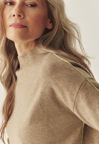 Béžový pletený sveter s vysokým golierom a jemnou rebrovanou textúrou, s kontrastným svetlozeleným akcentom pozdĺž švového spoja na ramene.