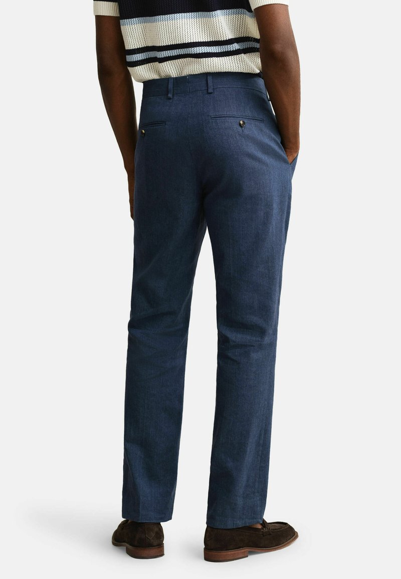 Uomo che indossa pantaloni su misura blu e mocassini scamosciati marrone scuro, con le mani in tasca, su uno sfondo chiaro e uniforme.