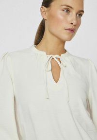 Blusa bianca con scollatura rotonda, chiusura a fiocco e maniche leggermente a sbuffo. Tessuto liscio con un leggero effetto lucido.