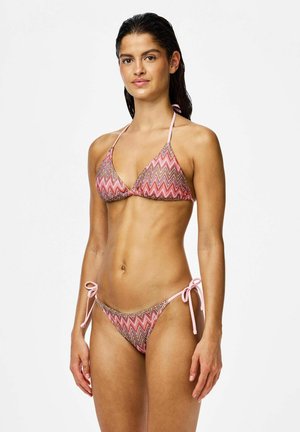 Donna in piedi che indossa un bikini rosa e marrone con motivo a zig zag e laccetti, su uno sfondo bianco semplice.