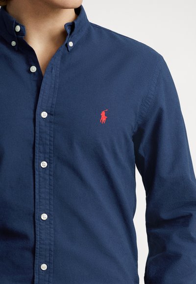 Navy blue button-down shirt in cotton blend, featuring a small red logo on the left chest and white buttons along the placket.