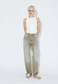 Jeans anchos de color gris claro con un acabado deslavado y bolsillos delanteros, combinados con una blusa blanca ajustada sin mangas y zapatillas claras.
