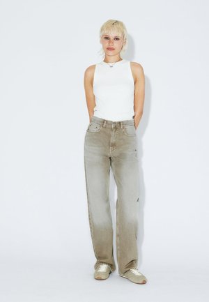 Young person with short blonde hair wearing white sleeveless top, loose beige jeans, and sneakers standing against a plain light background.