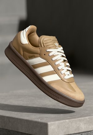 Beiger Adidas Samba Sneaker mit weißen Streifen, Schnürsenkeln und Fersendetail, schwebend über einer Steinoberfläche vor einem dunklen unscharfen Hintergrund.