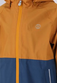 Vattentät jacka med en orange övre del och en blå nedre del. Inkluderar en framzip och en huva. Reflekterande logotypdetalj.