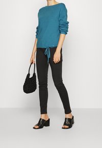 Un pull teal avec des manches retroussées et un ourlet noué, assorti à un jean skinny noir, des sandales à talons blocs ouvertes noire et un sac à main noir texturé.