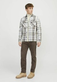 Geruite shirt in wit, beige en marineblauw met twee borstzakken, gecombineerd met een bruine broek en beige laarzen, staande tegen een lichte achtergrond.