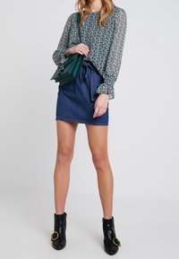 Blouse verte à motif floral avec manches longues bouffantes, associée à une mini-jupe en jean foncé et des bottines noires avec des accents dorés.