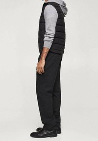 Schwarze Steppweste über einem grauen Hoodie, kombiniert mit dunklen Hosen und schwarzen Schuhen. Das Design ist figurbetont und die Texturen sind weich.