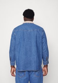 Jeansskjorta i blått med en beige krage, långa ärmar och knappmanschetter. Har en rak skärning och detaljer med bakåtkrafter.