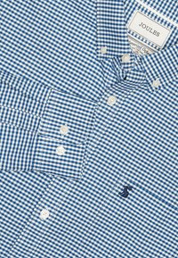 Niebiesko-biała koszula w kratę z długim rękawem, guzikami, kołnierzykiem na skład, oznakowana metką i haftowanym granatowym logo z zającem na piersi.