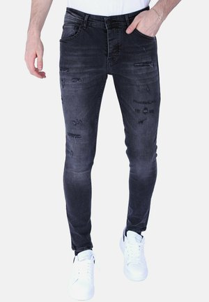 Man die zwarte slim-fit distressed jeans en witte sneakers draagt, met zijn hand in zijn zak, staand tegen een effen achtergrond.