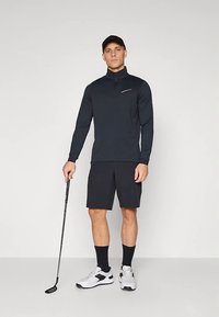 Melnā garā piedurkņu džemperis ar pus rāvējslēdzēju, kopā ar melniem šortiem. Rokā tur golfa nūju, un izskatu noslēdz baltas sporta kurpes.