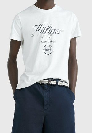 T-shirt en coton blanc avec un logo imprimé "Hilfiger New York" en gris foncé, à manches courtes, col rond, associé à un short marine et une ceinture tissée.
