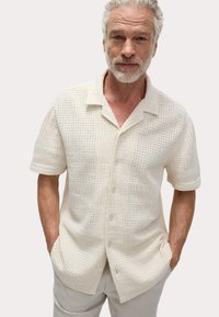 Chemise blanche à manches courtes avec un motif textur�é tricoté, fermeture à boutons sur le devant et coupe décontractée ; associée à un pantalon de couleur claire.